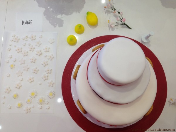 16. On ressort le premier gâteau du frigo, on le colle sur le socle avec un gros point de glucose liquide. Puis on fait de même avec le second gâteau. Oh ! C'est beau ! On termine en collant les fleurs et autres petites déco avec du glucose, on rajoute des déco en sucre si on veut (ici des citrons, pour rappeler les palets au citron du départ)... Et on admire !